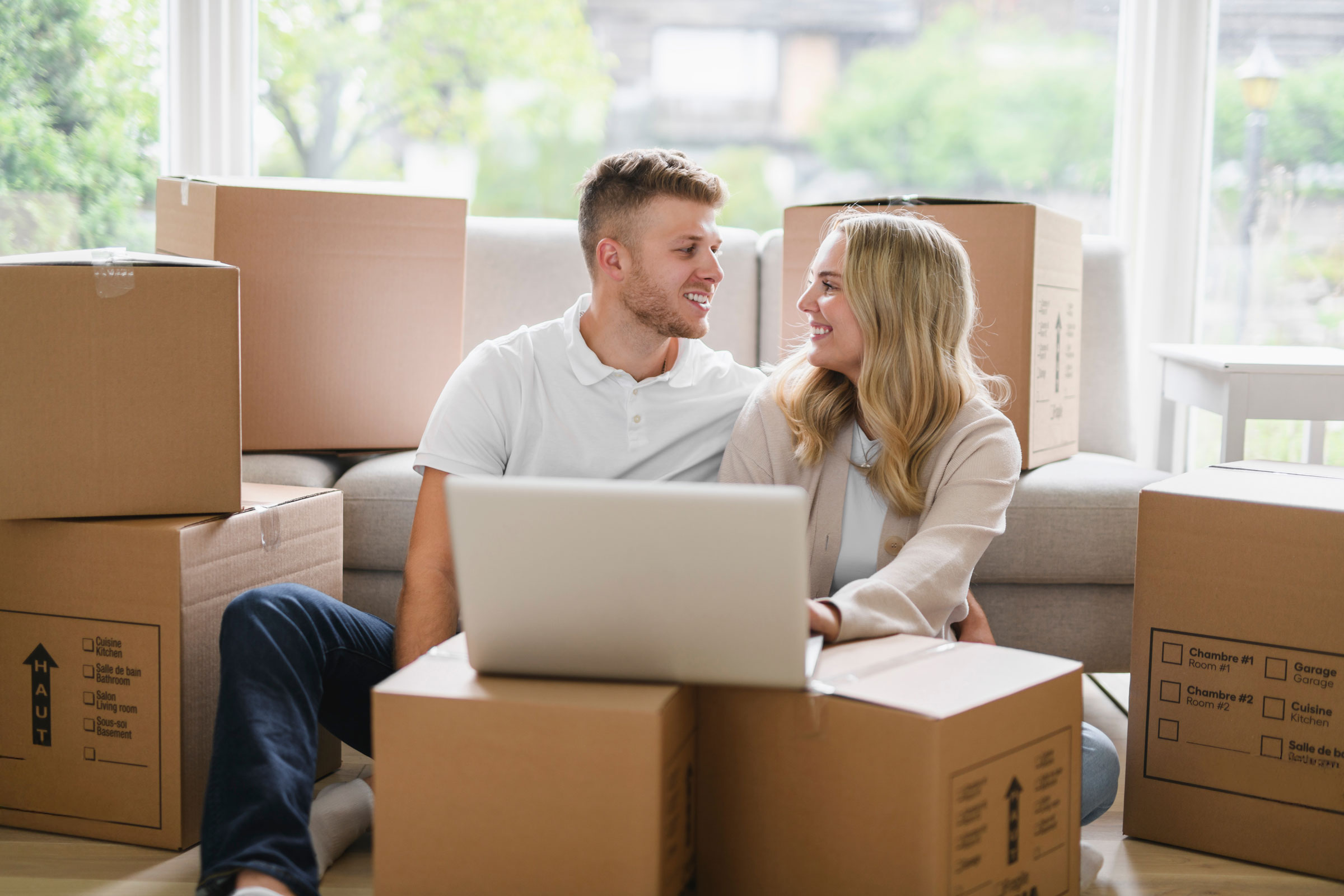 A couple sitting amongst a pile of boxes, using a laptop.