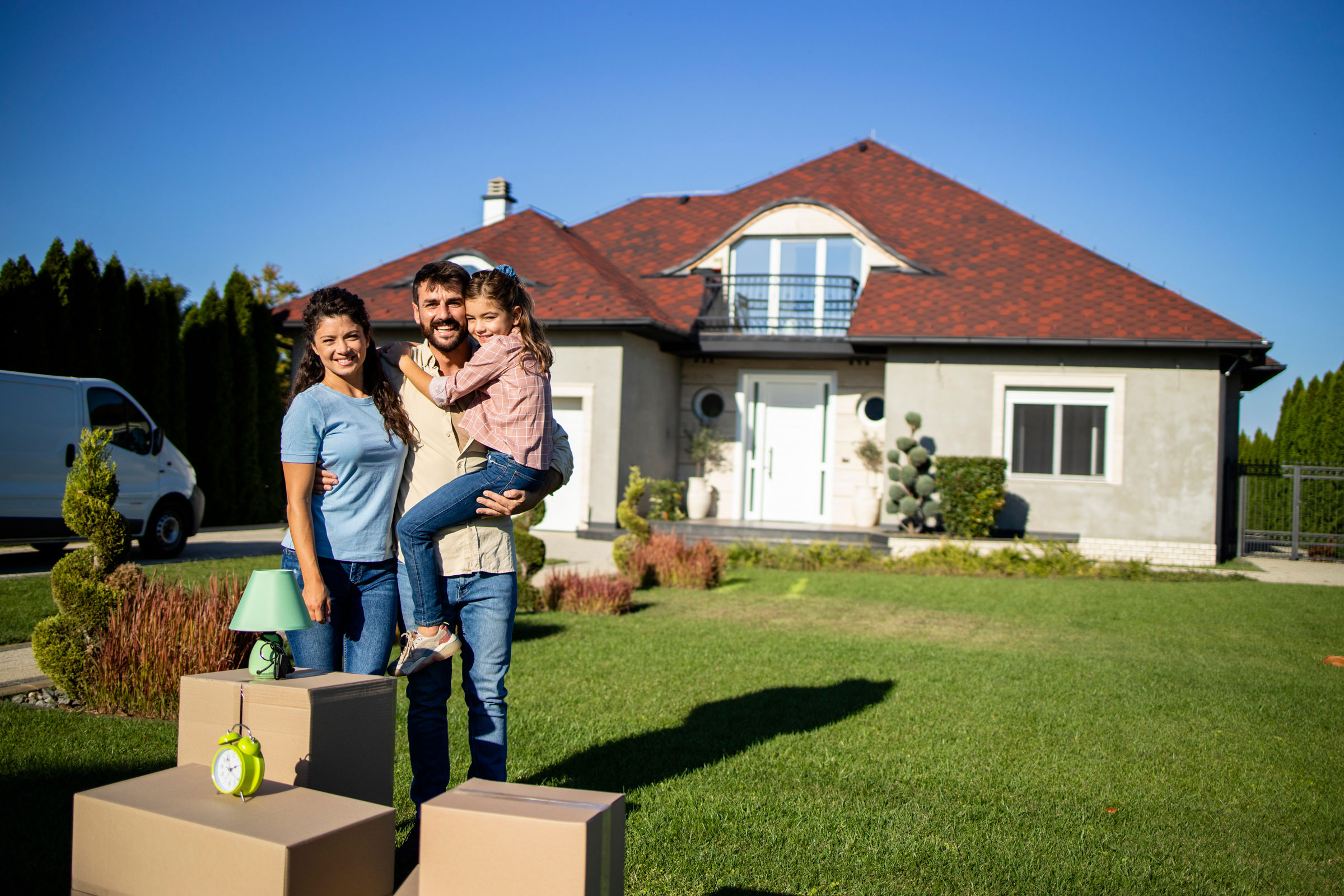 A couple with a child standing in front of their new house with packing boxes in front of them.